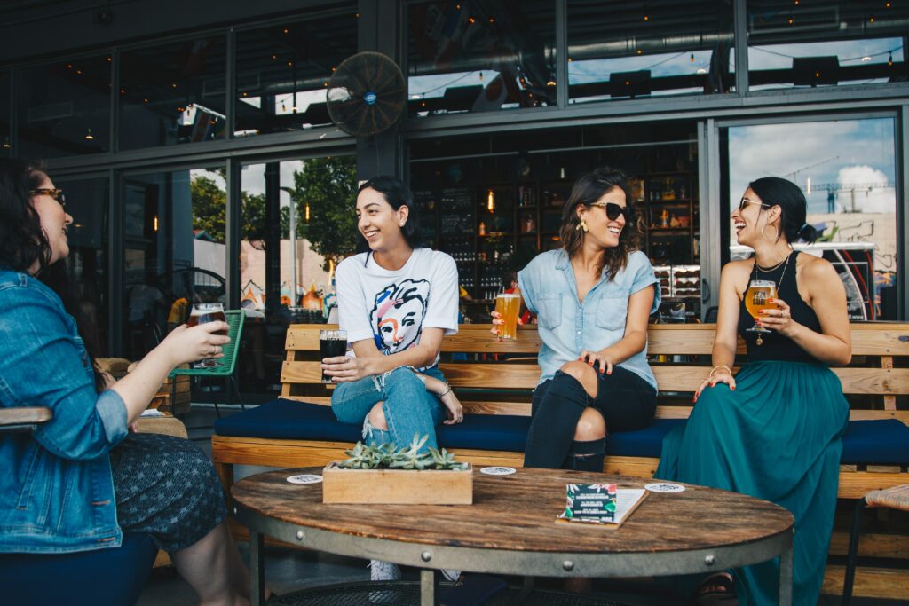 Four people sit on outdoor benches at a bar, laughing and holding drinks as they discuss marketing strategies. There are plants and coasters on the table in front of them, creating a vibrant networking atmosphere.