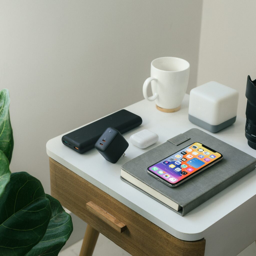 A smartphone, wireless earbuds, power bank, cube-shaped charger, notebook, mug, small lamp, and camera lens sit on a white desk next to a green plant—perfect for digital marketing or email marketing professionals.