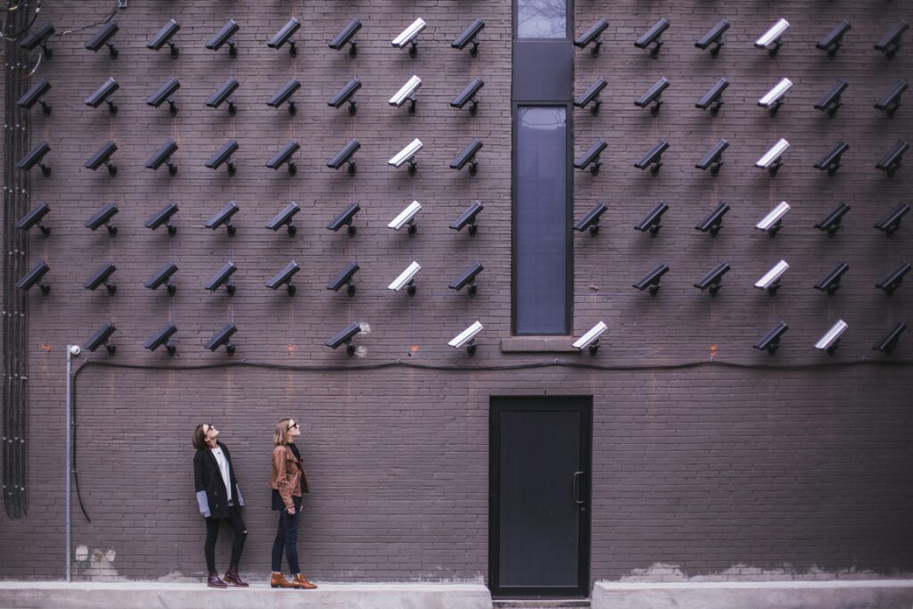 Two people stand in front of a brick wall covered with numerous black and white security cameras, arranged in rows—resembling the watchful strategies of search engine optimization and modern marketing.