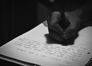 A person writes on a piece of graph paper with a pen. The hand is seen holding the pen, and the writing is in cursive, possibly outlining digital marketing or SEO strategies, though the text is not fully legible.
