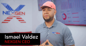 A man in a pink hat and gray polo shirt stands in an office with charts on screens behind him, highlighting NEXGEN CEO Ismael Valdez’s focus on search engine optimization and digital strategies.