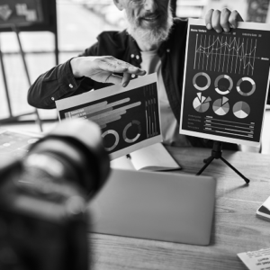 A person explains bar and pie charts related to digital marketing strategies in front of a camera during a presentation at a desk with a laptop and notebook.