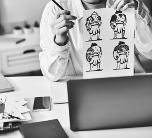 Person holding a sheet with four black and white cartoon-style illustrations of a character, seated at a desk with a laptop, smartphone, notebooks—perfect for brainstorming fresh marketing or SEO ideas.