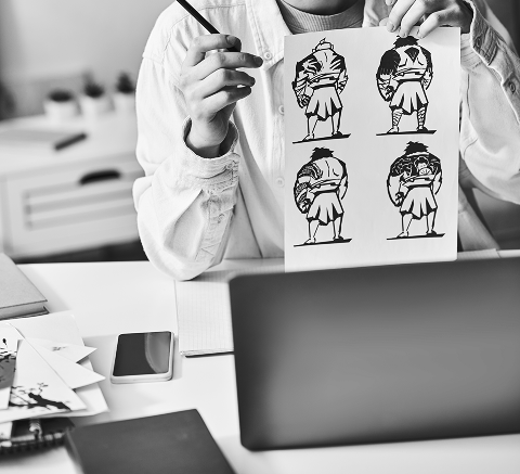 Person holding a sheet with four black and white cartoon-style illustrations of a character, seated at a desk with a laptop, smartphone, notebooks—perfect for brainstorming fresh marketing or SEO ideas.