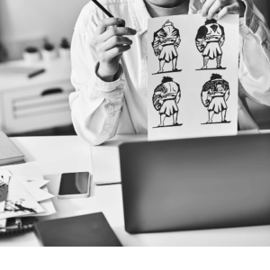 Person holding a sheet with four black-and-white character sketches at a desk, surrounded by tools for digital marketing—laptop, phone, notebook, and paper.