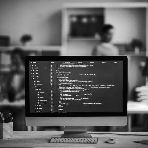 A computer monitor displays lines of code in a modern office environment, with two people blurred in the background, collaborating on marketing and SEO strategies.