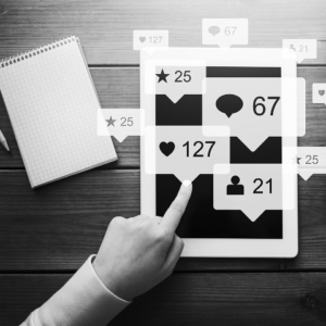 A hand touches a tablet displaying social media notifications—likes, comments, followers, and stars—on a wooden table with a notepad and pen nearby, highlighting the impact of digital marketing and CRM strategies.