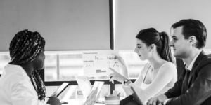 Three people sit at a table with laptops while one person holds up a chart and discusses digital marketing data during a meeting.