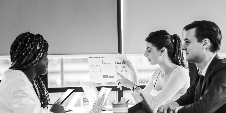 Three people sit at a table with laptops while one person holds up a chart and discusses digital marketing data during a meeting.