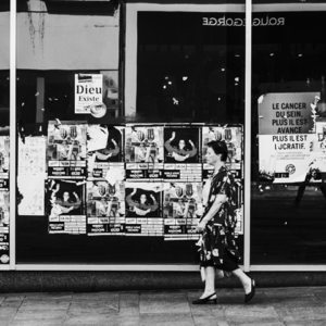 A woman in a patterned dress walks past a glass wall covered with marketing posters and signs, reflecting the street and buildings across from her. The image is in black and white.