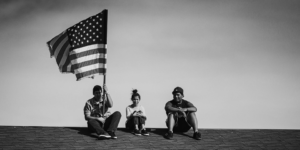 Three people sit on a rooftop; one holds an American flag. The black-and-white scene, framed by a clear sky, evokes the unity often celebrated in digital marketing campaigns.