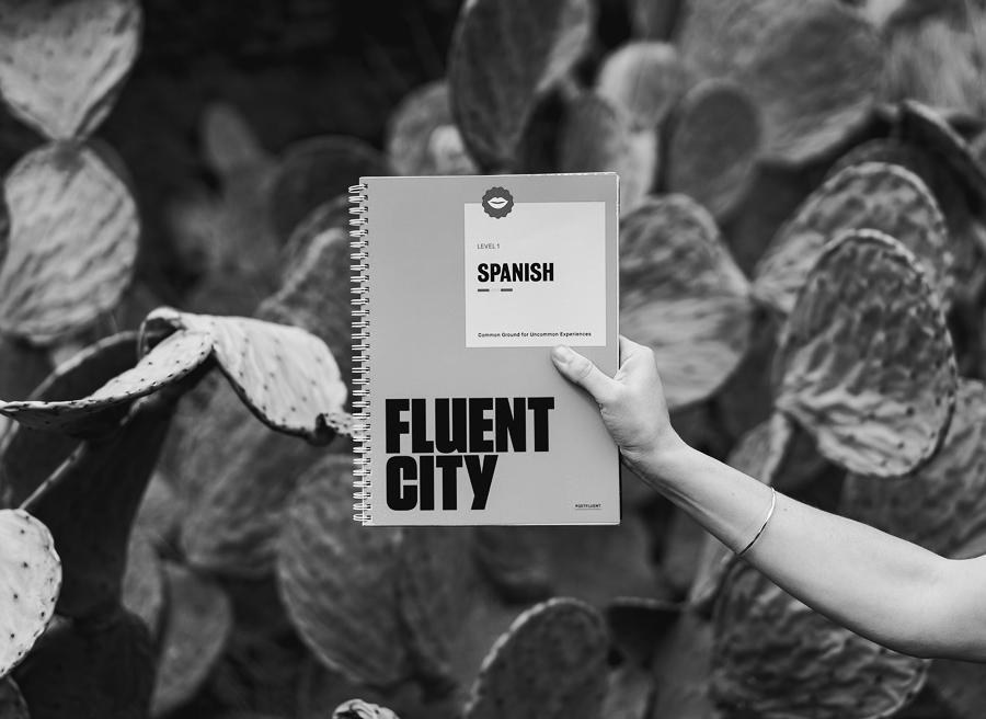 A person holds up a spiral-bound Spanish language workbook labeled "Fluent City" in front of large cactus plants, showcasing a unique marketing approach to language learning.