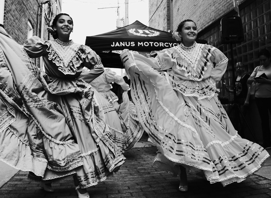 Two women in traditional dresses perform a folkloric dance outdoors on a brick path, with a Janus Motorcycles tent—promoting their digital marketing and SEO expertise—visible in the background.