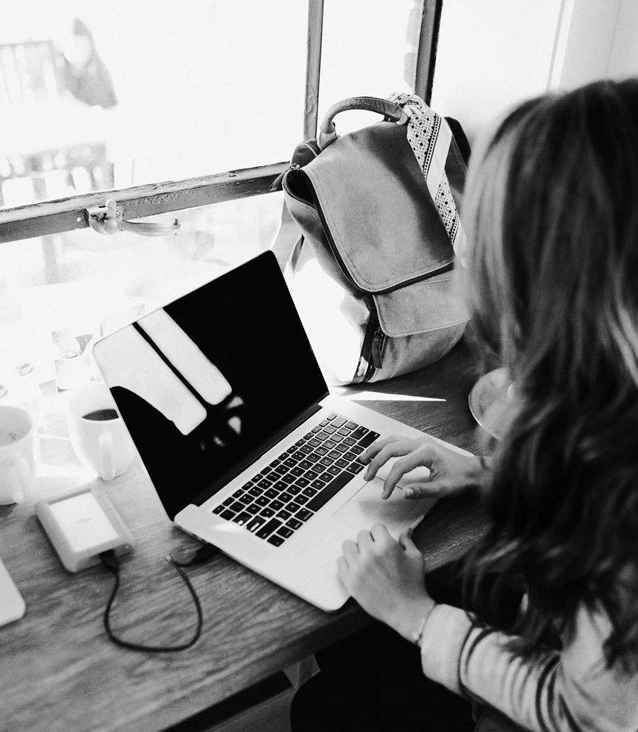 A person sits at a desk using a laptop, working on email marketing, with a backpack and external hard drive nearby, next to a window.