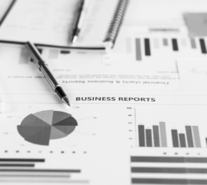 A pen rests on printed business reports featuring bar graphs, pie charts, and financial data related to digital marketing, with a spiral notebook in the background.