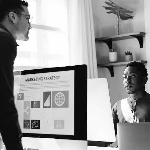 Two men in an office discuss a marketing strategy displayed on a computer screen, analyzing icons and graphs related to email marketing and campaign performance.