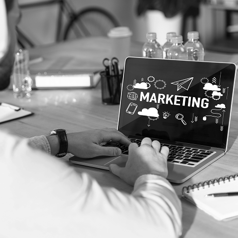 Person working on a laptop with the screen displaying "MARKETING" and related icons; water bottles and office supplies are on the table, highlighting a focus on digital marketing activities.