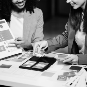 Two people in business attire review printed documents and charts at a table, discussing marketing strategies such as email marketing and search engine optimization while pointing at various papers and digital screens.
