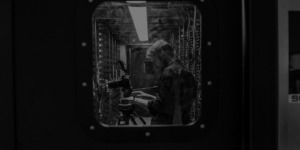 A person stands in a server room, adjusting digital marketing equipment next to metal racks, viewed through a window in a door.