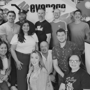A group of thirteen adults pose together indoors in front of a sign that partially reads "everage," perhaps hinting at crm or marketing. Most are smiling and facing the camera.