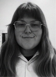 A person with long hair, glasses, and a septum piercing is facing the camera and smiling slightly. They are wearing a collared shirt, giving off a professional SEO or marketing specialist vibe.