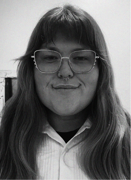 A person with long hair, glasses, and a septum piercing is facing the camera and smiling slightly. They are wearing a collared shirt, giving off a professional SEO or marketing specialist vibe.