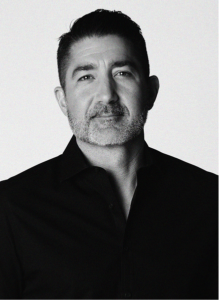 A man with short dark hair and a beard, dressed in a dark collared shirt, looks at the camera against a plain light background, projecting confidence often seen in digital marketing professionals.