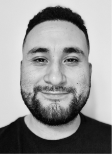 A man with short dark hair and a beard is smiling, facing the camera against a plain light background, embodying a friendly digital marketing professional.