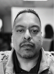 A man with short hair and a trimmed goatee looks directly at the camera in a black-and-white photo, with blurred people and objects in the background—capturing focus much like effective email marketing.