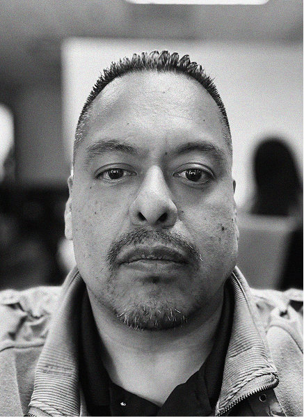 A man with short hair and a trimmed goatee looks directly at the camera in a black-and-white photo, with blurred people and objects in the background—capturing focus much like effective email marketing.