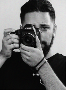 Man with a beard holds a Canon DSLR camera up to his face, looking directly into the lens, against a plain background—capturing the focus much like effective SEO draws attention in search engine optimization.