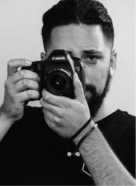 Man with a beard holds a Canon DSLR camera up to his face, looking directly into the lens, against a plain background—capturing the focus much like effective SEO draws attention in search engine optimization.