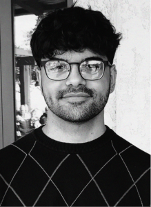 A person with short, dark hair, glasses, and a trimmed beard wears a dark, diamond-patterned sweater, standing in front of a textured wall—poised and professional, perfect for any digital marketing or search engine optimization team.