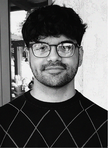 A person with short, dark hair, glasses, and a trimmed beard wears a dark, diamond-patterned sweater, standing in front of a textured wall—poised and professional, perfect for any digital marketing or search engine optimization team.