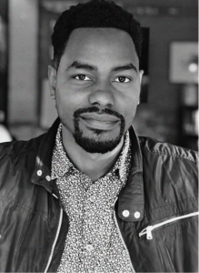 A man with short curly hair and a trimmed beard is wearing a patterned shirt and a zippered jacket, looking directly at the camera in a well-lit indoor setting, embodying a professional vibe often seen in digital marketing or SEO profiles.