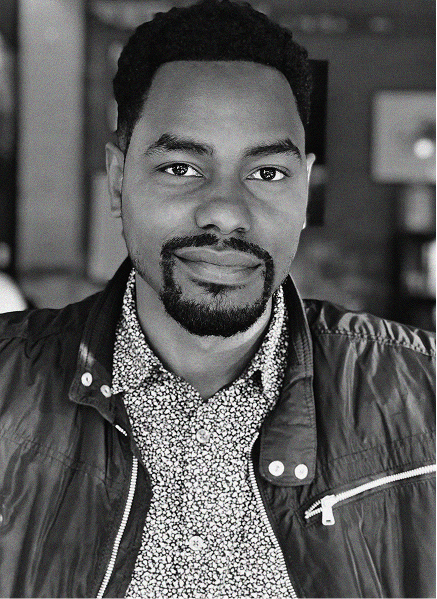 A man with short curly hair and a trimmed beard is wearing a patterned shirt and a zippered jacket, looking directly at the camera in a well-lit indoor setting, embodying a professional vibe often seen in digital marketing or SEO profiles.