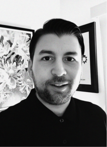 A man with short dark hair and a trimmed beard, wearing a dark collared shirt, stands indoors in front of framed wall art. The black and white image reflects the precision often sought in SEO and search engine optimization strategies.