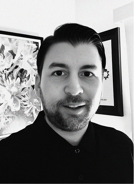 A man with short dark hair and a trimmed beard, wearing a dark collared shirt, stands indoors in front of framed wall art. The black and white image reflects the precision often sought in SEO and search engine optimization strategies.