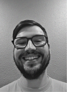 A person with glasses and a beard, perhaps a digital marketing expert, smiles at the camera in front of a textured wall. The image is in black and white.