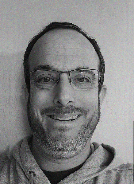 A man with glasses, a short beard, and a hoodie smiles at the camera against a plain, light-colored wall, embodying the modern spirit of digital marketing.