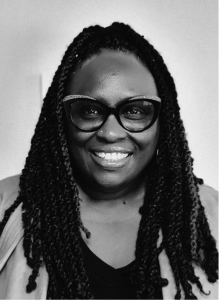 A woman with long braided hair, wearing dark-rimmed glasses and a dark top, smiles at the camera against a plain background, reflecting confidence in digital marketing.
