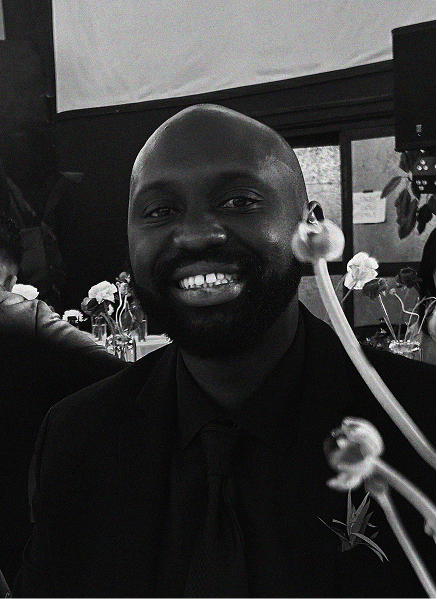 A man in a suit smiles at the camera in a black and white photo, with blurred decorative elements and flowers in the foreground—reflecting the polished image often featured in digital marketing campaigns.