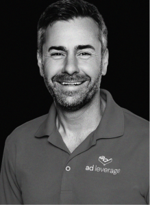 A man with short hair and a beard smiles, wearing a polo shirt with the "ad leverage" logo, representing expertise in marketing, against a black background.