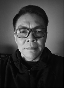A man wearing glasses and a dark zip-up jacket looks directly at the camera in a black and white portrait, capturing the composed confidence of a digital marketing professional.