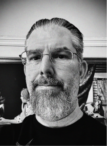 Black and white close-up photo of a man with glasses and a beard, standing indoors in front of a wall with framed pictures and some marketing figurines visible in the background.