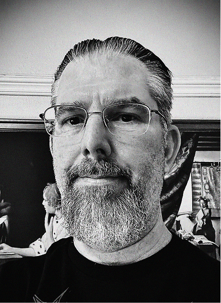 Black and white close-up photo of a man with glasses and a beard, standing indoors in front of a wall with framed pictures and some marketing figurines visible in the background.