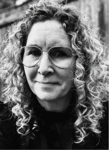 Black and white portrait of a person with curly hair and large glasses, looking at the camera with a slight smile. A wooden fence is visible in the blurred background, evoking a thoughtful mood perfect for marketing or CRM campaigns.