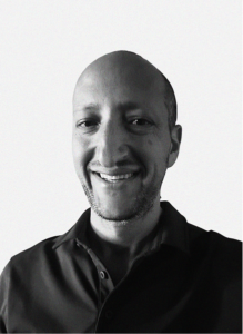 A man with a shaved head and short beard smiles at the camera while wearing a dark collared shirt against a plain background, reflecting confidence often seen in digital marketing professionals.