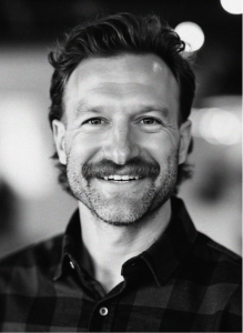 A man with a mustache and beard smiles at the camera, wearing a checkered shirt. The blurred background hints at a creative marketing or digital marketing workspace.