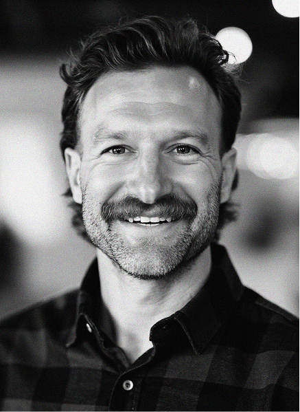 A man with a mustache and beard smiles at the camera, wearing a checkered shirt. The blurred background hints at a creative marketing or digital marketing workspace.